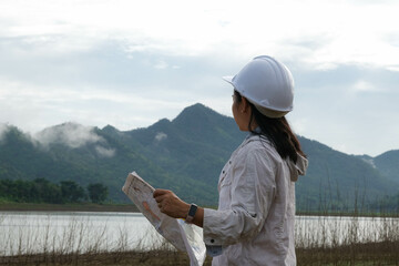 Engineering ecologist woman in a helmet holding a blueprint stands on the bank of a river to...