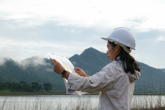 Engineering ecologist woman in a helmet holding a blueprint stands on the bank of a river to develop a hydroelectric dam to generate electricity. Clean energy and Technology concepts.
