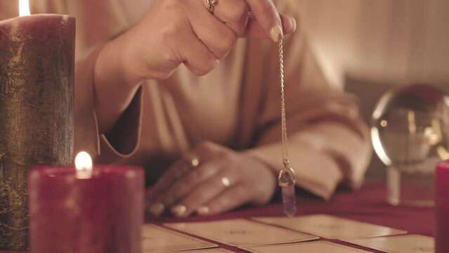 Cropped slowmo closeup of unrecognizable jeweled fortune teller holding pendulum over tarot cards spread on table