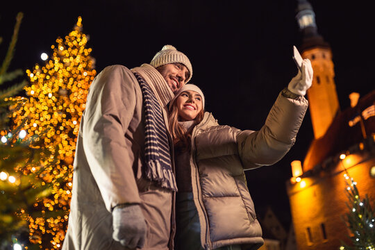 winter holidays and people concept - happy smiling couple taking selfie with smartphone over christmas tree lights in evening city