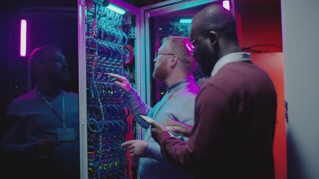 Caucasian data center technician checking cables in server room and speaking to African American colleague as he using digital tablet