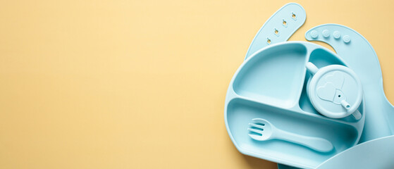 Flat lay silicone baby plate, cutlery, bib on pastel yellow background. Baby tableware set top view, copy space.