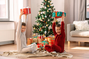 Obraz na płótnie Canvas christmas, winter holidays and childhood concept - happy girl and boy in pajamas with gifts sitting on floor at home