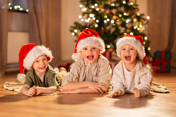 Fototapeta na wymiar christmas, winter holidays and childhood concept - happy little girl and boys in santa hats lying on floor at home