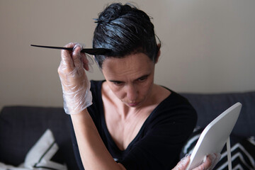 Middle age woman colouring dark hair with gray roots at home. Woman sitting on couch and dyeing hair using black brush and looking to the mirrow