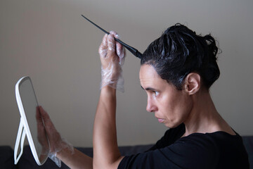 Middle age woman colouring dark hair with gray roots at home. Woman sitting on couch and dyeing hair using black brush and looking to the mirrow