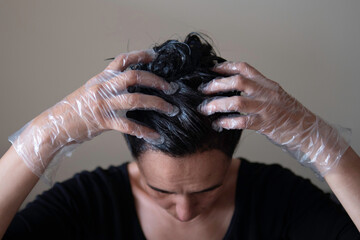 Naklejka premium Middle age woman colouring dark hair with gray roots at home. Woman sitting on couch and dyeing hair using black brush and looking to the mirrow