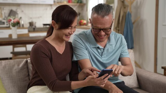 slow motion of happy asian elderly dad and adult daughter enjoying funny online video on smartphone together in the living room at home.
