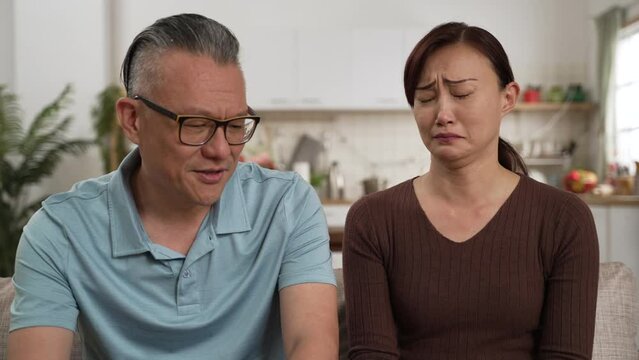 caring asian retired father consoling frustrated daughter and patting on her back in living room at home. the depressed woman cries and buries her face in hands while listening