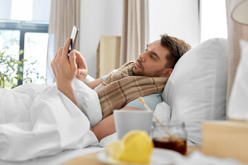health, illness and people concept - sick man with smartphone in bed at home