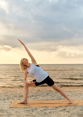 fitness, sport, and healthy lifestyle concept - woman doing yoga triangle pose on beach over sunset