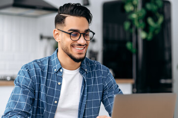 Close-up photo of excited positive indian or arabian guy with glasses, freelancer or creative designer, sitting at a desk at home, working on a creative project uses laptop, looks at screen, smiles