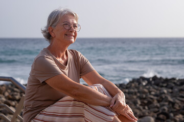 Fototapeta na wymiar Portrait of beautiful senior woman sitting at the beach at sunset looking away smiling - elderly relaxed lady enjoys vacation and freedom