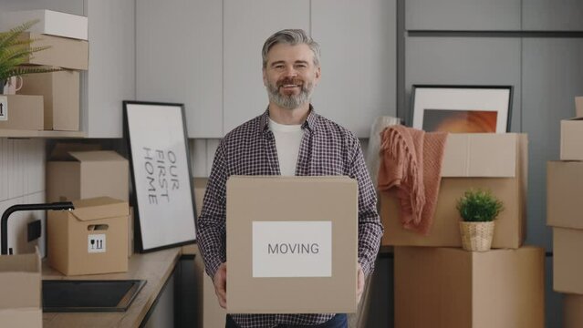 Portrait of middle aged man owner with boxes standing at new home. Cheerful husband caring paper boxe in slow motion. New home and moving concept.