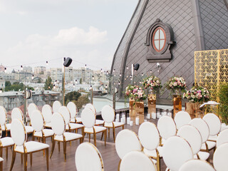 The decor of the outdoor wedding is on the roof against the backdrop of a classic brown tower with an oval window. Gold decor, white chairs, bouquets of flowers, candles on the floor, garlands.