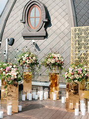 The decor of an outdoor wedding on the roof against the backdrop of a brown tower with a window. Bouquets of white and pink flowers stand on gold stands and white candles in candlesticks on the floor.