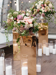 An exotic bouquet of protea, dry hydrangea, white and cream roses, barberry sprigs, and leaves stands in a glass vase on a golden stand. Floral decor and white candles in the background.