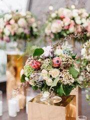 An exotic bouquet of protea, dry hydrangea, white and cream roses, barberry sprigs, and leaves stands in a glass vase on a golden stand. Floral decor and white candles in the background.