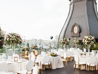 The decor of the wedding banquet is white on the roof against the backdrop of a brown tower with an oval window. On the table is a white tablecloth, cutlery, plates, wine glasses, bouquets of flowers.