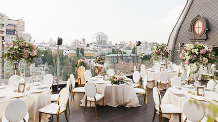 Fototapeta na wymiar The decor of the wedding banquet is white on the roof against the backdrop of a brown tower with an oval window. On the table is a white tablecloth, cutlery, plates, wine glasses, bouquets of flowers.