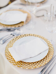 Banquet table setting in gold and white tones. Golden and white plate, wine glasses, cutlery, white napkin on a plate on a white tablecloth.