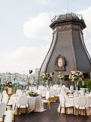 The decor of the wedding banquet is white on the roof against the backdrop of a brown tower with an oval window. On the table is a white tablecloth, cutlery, plates, wine glasses, bouquets of flowers.