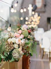 Floral decor for a party on the terrace in the open air. In the foreground is a bouquet of green hydrangea, white and cream roses, peonies, and green leaves. In the background are glowing garlands.