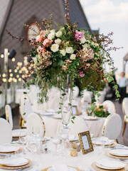 Wedding table setting in gold and white. On the table are a white tablecloth, dishes, cutlery, and a bouquet of flowers in a tall glass vase. In the background is a brown tower with an oval window.