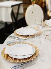 Banquet table setting in gold and white tones. Golden and white plate, wine glasses, cutlery, white napkin on a plate on a white tablecloth.