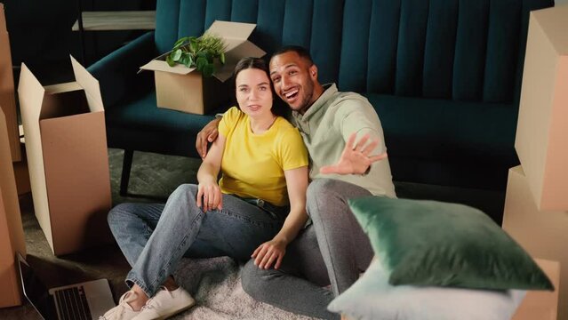 Happy interracial couple talk, sitting on a living room of their new home with a lot of boxes in the background. Married couple plan apartment decoration and imagine their beautiful life. 