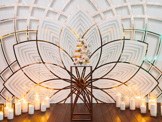 A four-tiered white wedding cake with golden ribbons, decorated with flowers, stands on a golden table against the backdrop of a lotus. There are white burning candles on the floor.