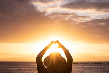 Back view of woman silhouette admiring the orange sunset over the sea doing heart shape with her...