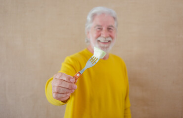 Defocused white-haired senior man in yellow shirt isolated on a light background holds a fork with a piece of melon ready to be eaten