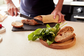 Woman hands is cutting a French baguette in the kitchen. Basil and whole grain cutting bread. Selective focus. Blurred background