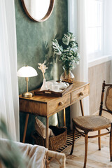 Console table with a table lamp, vase with live plants and a vintage wooden chair in a room with green textured walls, large windows with curtains and a wooden floor. Empty space
