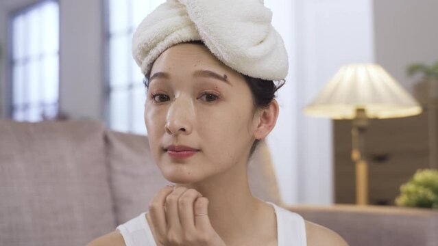 close up asian girl looking at camera as mirror with towel on head is examining her smooth skin and smiling with satisfaction at cozy living room background.