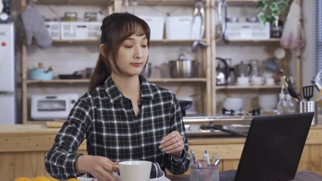 beautiful asian girl carrying tea is returning to her seat at dining table and putting on the headphone to continue with school online lesson at home.