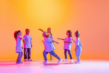 Naklejka premium Portrait of cheerful, active little girls, happy kids dancing isolated on orange background in neon light. Concept of music, fashion, art, childhood, hobby