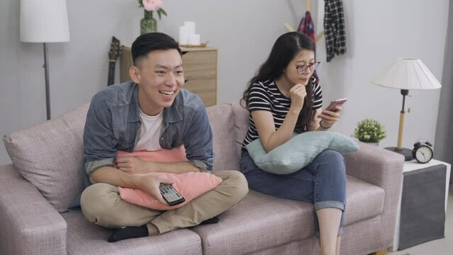 portrait lady is sitting phubbing while her boyfriend watching tv next to her is turning up the volume with a remote control on the sofa at home.