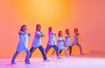 Portrait of cheerful, active little girls, happy kids dancing isolated on orange background in neon light. Concept of music, fashion, art, childhood, hobby