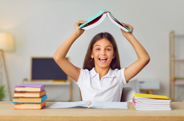 Portrait of funny schoolgirl sitting at desk and holding open book above her head and laughing. Cute preteen girl sitting between books and coopybooks in classroom or at home. Back to school concept.