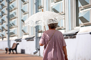 Old woman with sun umbrella walk on a street. Hot weather, life of elderly people in summer