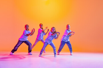 Group of children, little girls in sportive casual style clothes dancing in choreography class isolated on orange background in purple neon light.