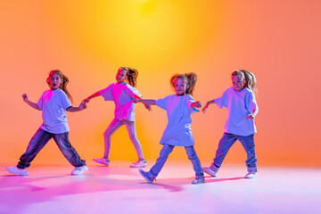 Hip-hop dance, street style. Happy children, little active girls in casual style clothes dancing isolated on orange background in purple neon light.
