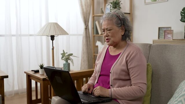 asian old lady is gesturing hi and talking touching her chest while having a online consultation through video call on the computer in the living room at home