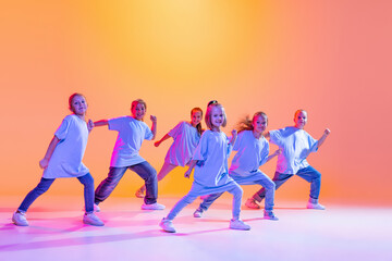 Hip-hop dance, street style. Happy children, little active girls in casual style clothes dancing isolated on orange background in purple neon light.