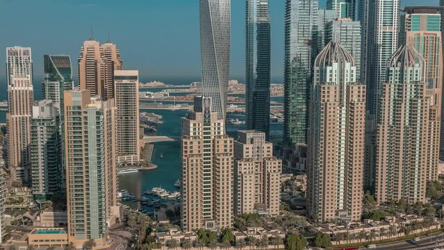 Skyscrapers of Dubai Marina with illuminated highest residential buildings morning timelapse. Boats floating in canal. Aerial top view from JLT district during sunrise with warm light