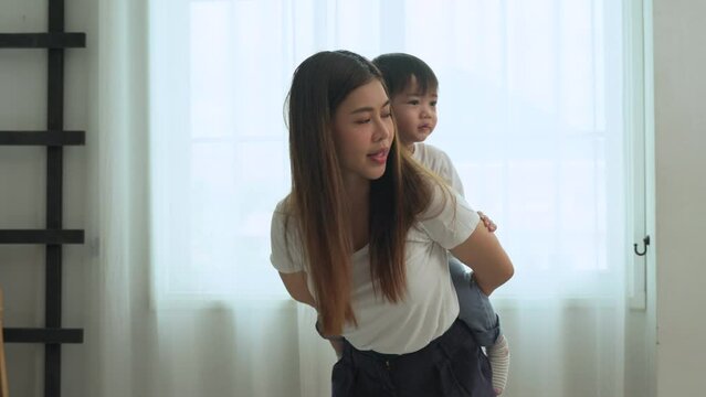 Asian young mother lulling a lovely son while giving piggyback ride to her little son in the living room at house.