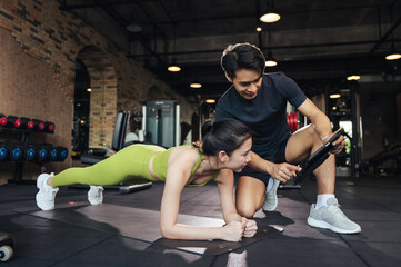 Fototapeta na wymiar Personal trainer looking at digital tablet and explaining progress to young woman at the fitness gym. Asian man trainer coach helping young female planking exercise for beaultiful building slim body.
