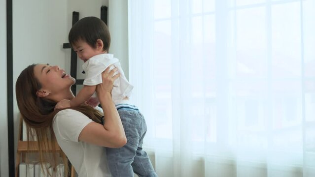 Asian young mother kissing and throwing her little son affectionately in the living room at house.